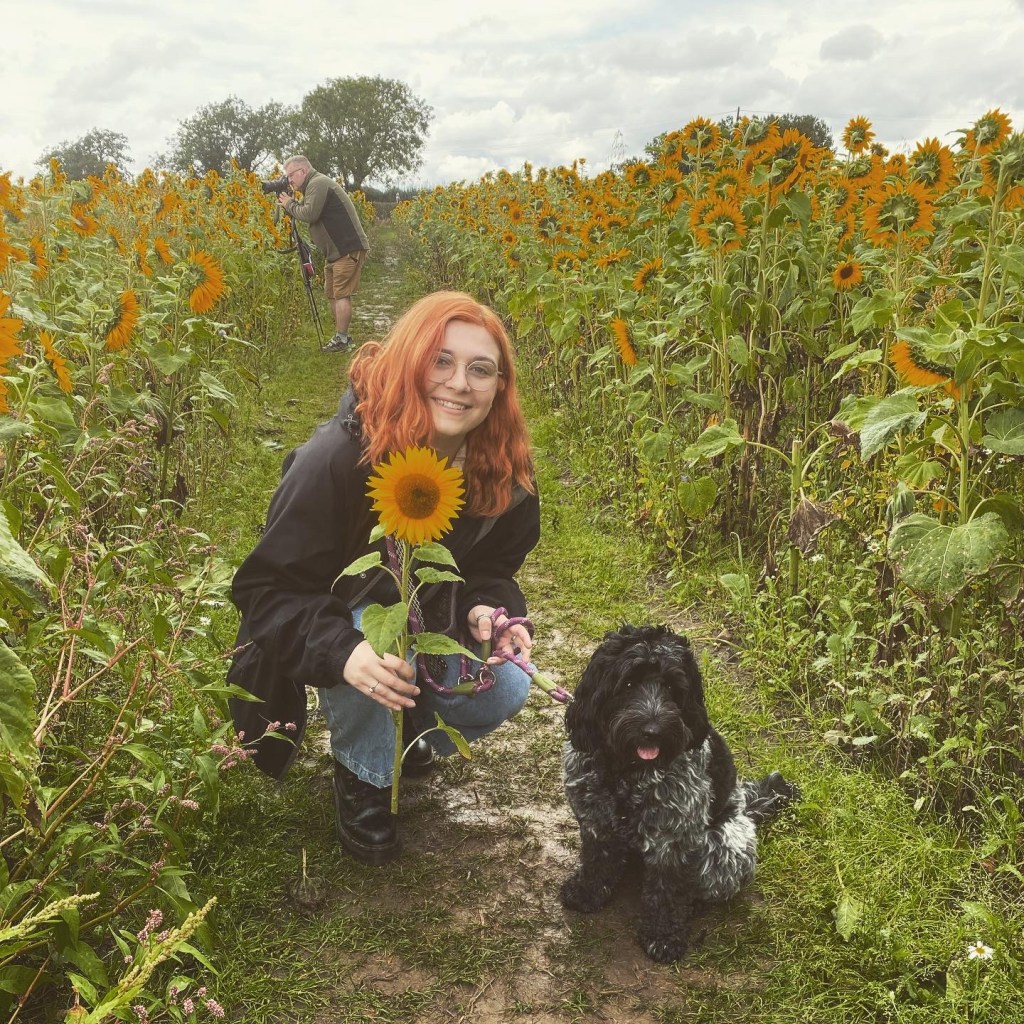 Beth, in a sunflower field, holding a sunflower and the lead of Wilson, a little black and white cockapoo. The sunflowers match Beth's hair, which is currently a peach-y colour.